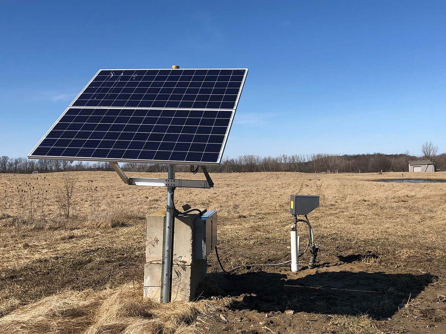 A solar panel mounted on a metal pole in an open field with dry grass. The panel is angled towards the sky, and a small control box is connected by wires near the base. In the background, there are leafless trees and a clear blue sky, hinting at an installation possibly supporting a top-head drive pump.