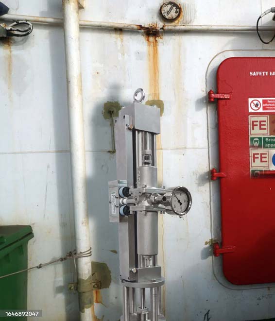 A vertical metal apparatus with gauges and valves, potentially part of a top-head drive pump system, is mounted on a wall with rust stains. To the right, there is a red safety door with warning signs. The surroundings appear industrial, with pipes and a green box on the left.