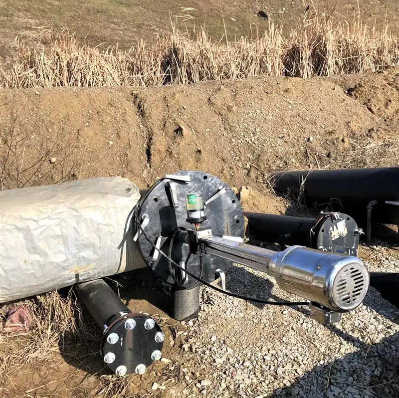 A large metal pipe with an attached motorized top-head drive pump is partially buried in dirt and gravel outdoors. The pipe is connected to several smaller black pipes. Dry grass and a dirt embankment are visible in the background.