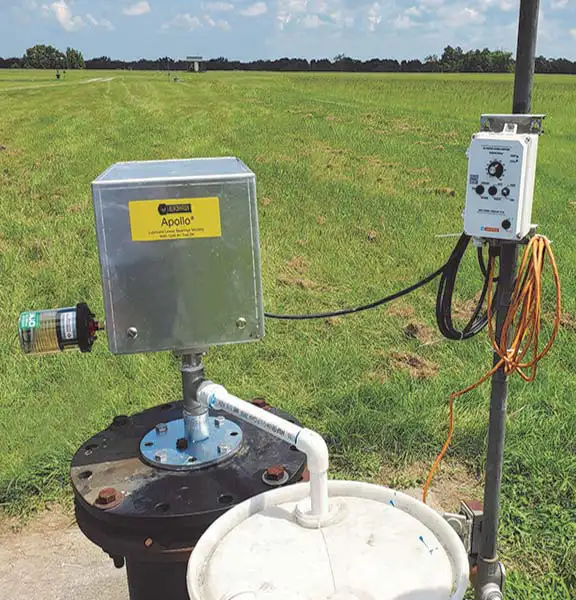 A metal box labeled "Apollo" is attached to a pipe structure in a grassy field, seemingly part of a top-head drive pump system. An electrical control device is affixed to a pole nearby, connected by wires and an orange extension cord. The sky is clear with scattered clouds in the distance.