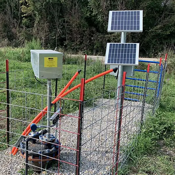 A fenced area containing a solar-powered device with two solar panels mounted on a pole and several pipes connected to a top-head drive pump system on the ground, surrounded by gravel. The setup is situated in a green, grassy outdoor area with trees in the background.