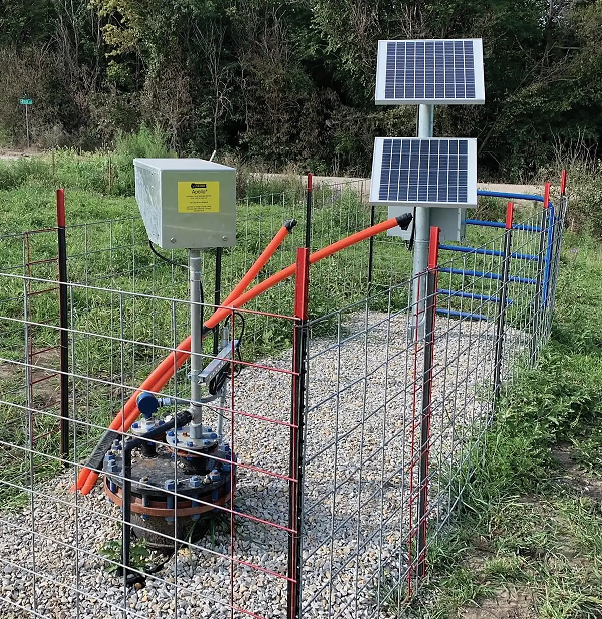 A fenced-off water well with orange pipes connected to a control box and blue pumps, including a top-head drive pump. Positioned nearby are two solar panels mounted on metal poles. The setup is situated outdoors, surrounded by grass and trees. Gravel surrounds the well base, with another fence in the background.