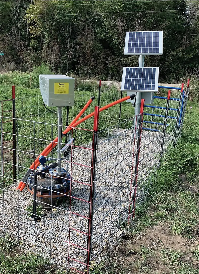 A solar-powered water pump system, featuring a top-head drive pump, is enclosed in a metal wire fence. Solar panels are mounted on posts alongside a control box and various pipes and valves. The area surrounding the pump is covered with gravel, bordered by grass and trees.