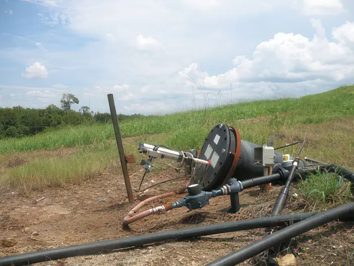 An outdoor machinery setup is placed on a dirt patch, surrounded by various pipes and cables. The machinery, featuring a large circular component and a top-head drive pump, appears to be related to drilling or irrigation. It is situated in a grassy area with trees and a cloudy sky in the background.