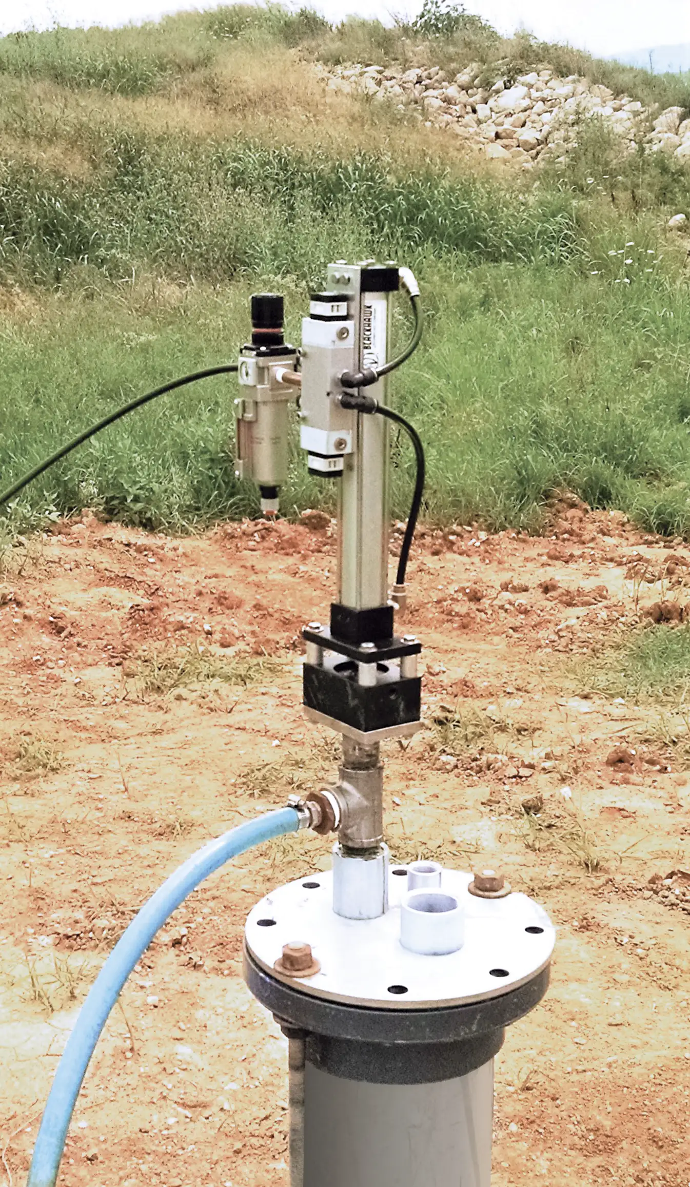 A top-head drive pump is mounted on a cylindrical structure with hoses connected, located outdoors on a dirt patch with grassy surroundings and rocks in the background.