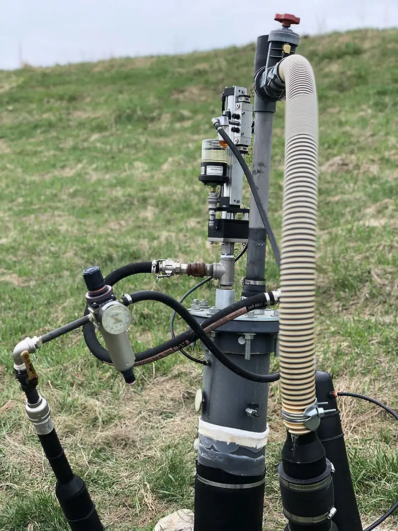 A close-up view of a well water monitoring system in a grassy outdoor area. The setup includes various pipes, hoses, a top-head drive pump, a gauge, and a valve with a red handle. The equipment is connected to a vertical metal pipe and surrounded by other mechanical components.