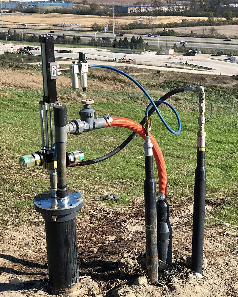 An outdoor setup featuring various pipes, valves, and gauges connected to a gas extraction wellhead with a top-head drive pump. The equipment is installed on a grassy hill with a highway and industrial buildings visible in the background. Colorful hoses are attached to the apparatus.