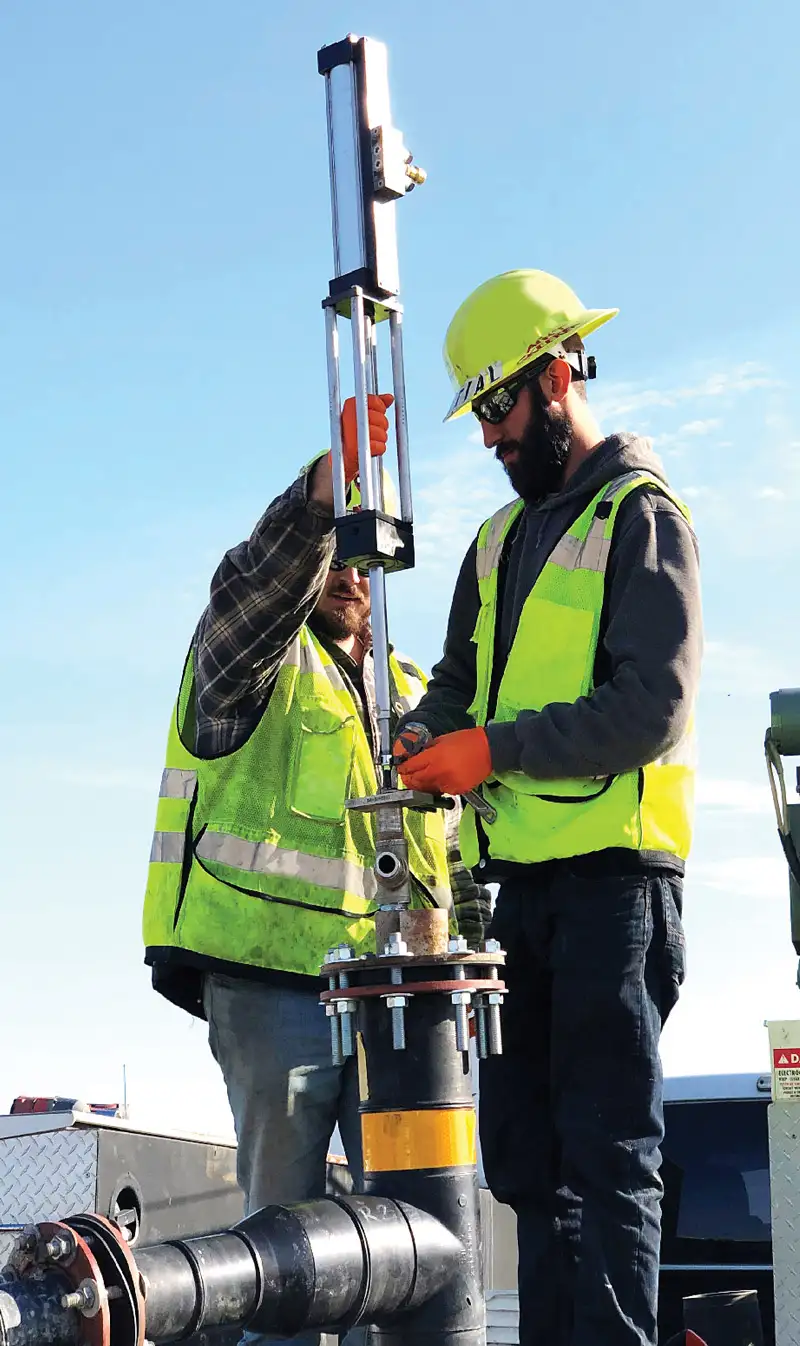 Two workers wearing yellow safety vests and hard hats adjust equipment on a pipe. One worker, in a flannel shirt, holds a tall instrument attached to the top-head drive pump, while the other, in a hoodie, works on a connection. The sky is clear and blue in the background.
