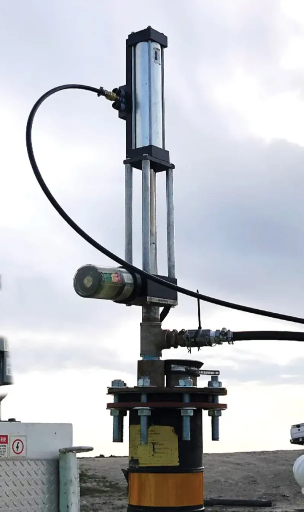 Close-up image of a mechanical industrial device with cylinders, pipes, and cables against a cloudy sky background. The equipment, featuring a top-head drive pump, is secured with large bolts and seems to be part of a larger setup.