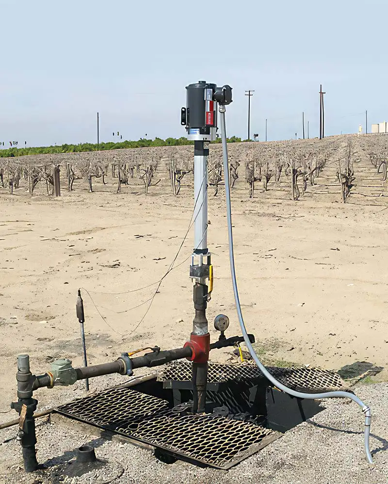 An image of a top-head drive pump installed in a dry agricultural field, surrounded by barren rows of vines or trees. The pump has metal pipes extending horizontally and is fixed on a grated platform. Nearby, electrical and control equipment is seen attached to the main vertical pipe.