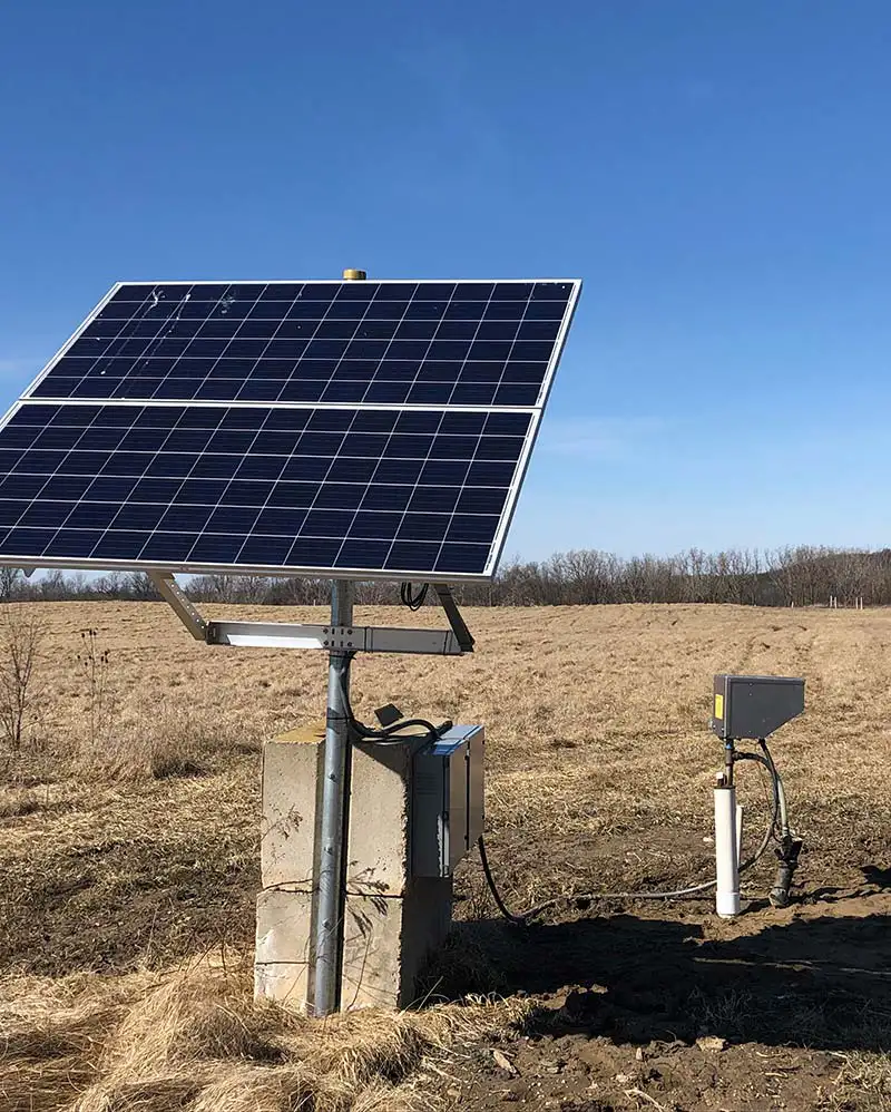 A solar panel is mounted on a metal stand in the middle of a field, powering a top-head drive pump. The panel is connected to a control box and a small white pipe. The sky is clear and blue, and the surrounding area is covered with dry grass.