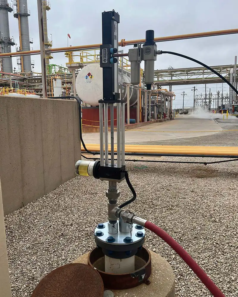 A top-head drive pump is installed in an industrial setting, with a grey control unit mounted atop a vertical metal structure. A red hose is connected to the pump, leading away from the structure. Pipes and industrial structures are visible in the background.