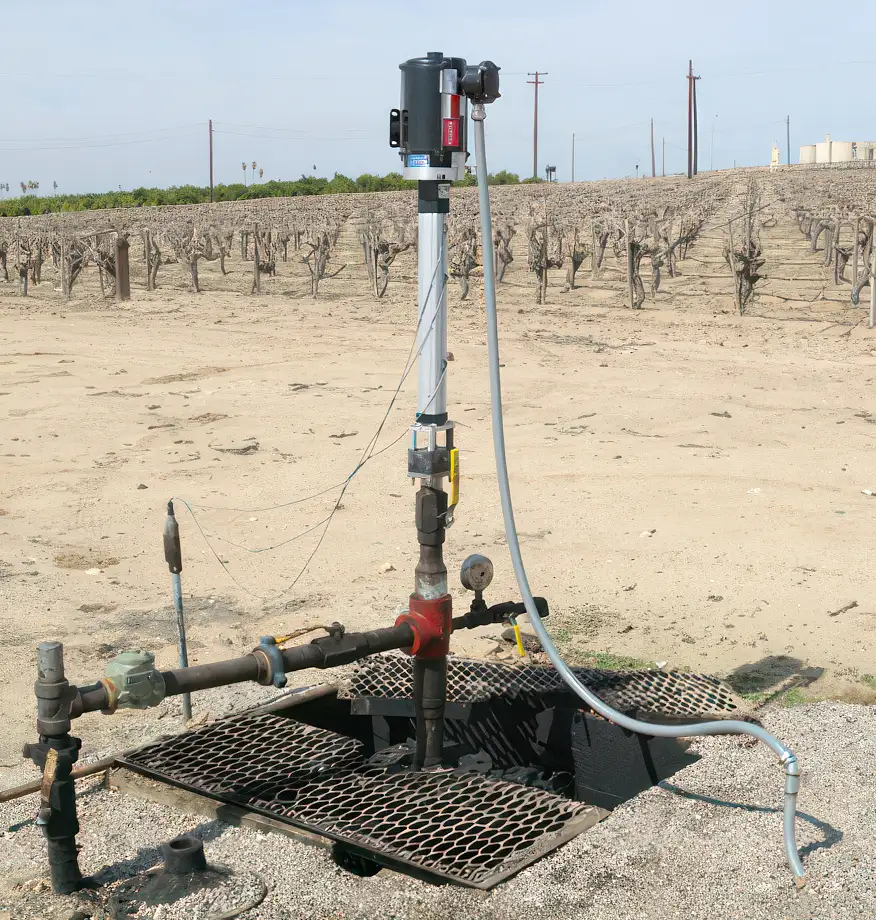 A well pump setup in an agricultural field features a top-head drive pump installed above a metal grate covering the well. Piping and electrical connections are attached to the vertical pump. In the background, barren rows of vines indicate a vineyard in its dormant season.