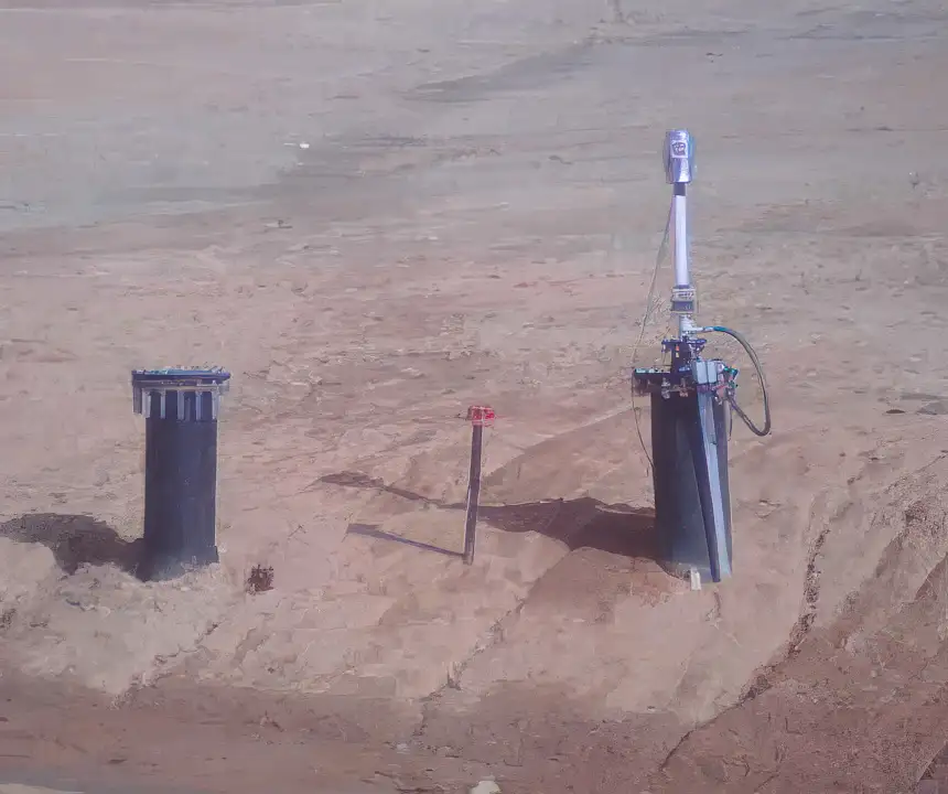 Three pipes of varying sizes and designs emerge from a barren, sandy landscape. Two larger pipes are partially buried, and the third, smaller pipe is also surrounded by sand. One large pipe has cables connected to it, resembling a top-head drive pump setup. The scene appears desolate and lacks vegetation.