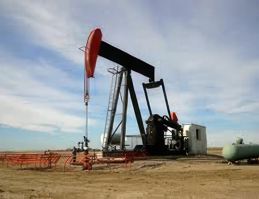 An oil pumpjack with a top-head drive pump stands on a barren piece of land under a partly cloudy sky. The structure is black with a red counterweight, and it is connected to machinery on the ground. A small building and cylindrical tank are also visible.
