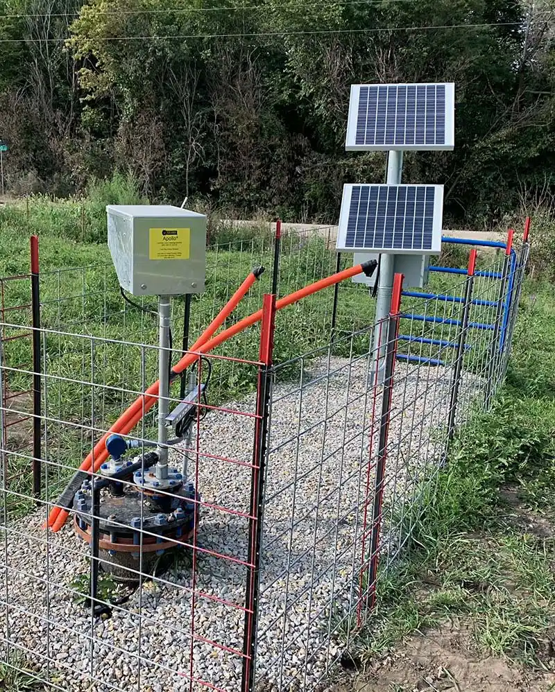 A fenced area with a solar-powered water pump system, featuring a top-head drive pump. The setup includes two solar panels mounted on a pole, red pipes, and control equipment encased in a grey box. Surrounding the equipment is a gravel path with green vegetation in the background.
