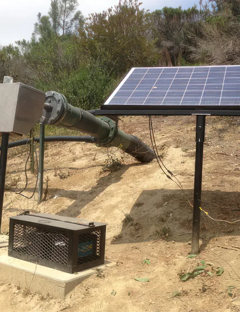 A solar panel is mounted on a metallic frame on a dirt hill. Nearby, a large, green pipe partially buried in the ground is connected to a top-head drive pump in a metal box. A smaller metal cage-like structure on the ground houses electrical equipment.