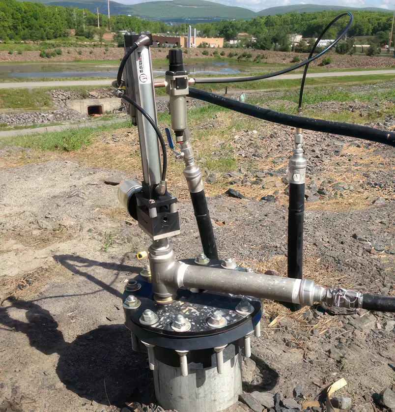 Close-up view of a groundwater monitoring well installation, featuring a well cap with various pipes, valves, and sensors above ground, including a top-head drive pump. The background includes a grassy landscape, trees, and distant buildings under a clear blue sky.