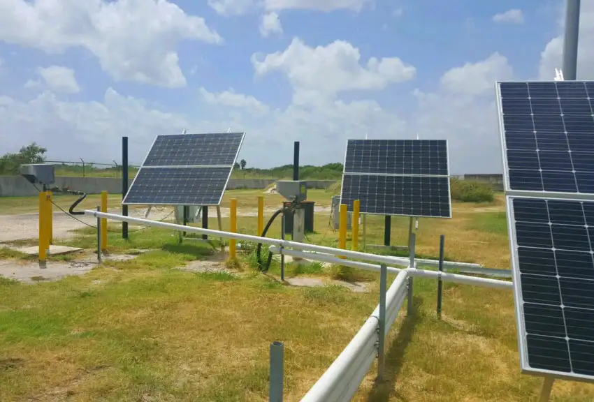 Three solar panels mounted on yellow and black stands are positioned in an open grassy area. Near the setup, a top-head drive pump is connected with wiring and pipes that run to a central unit. The sky is partly cloudy with blue patches visible.