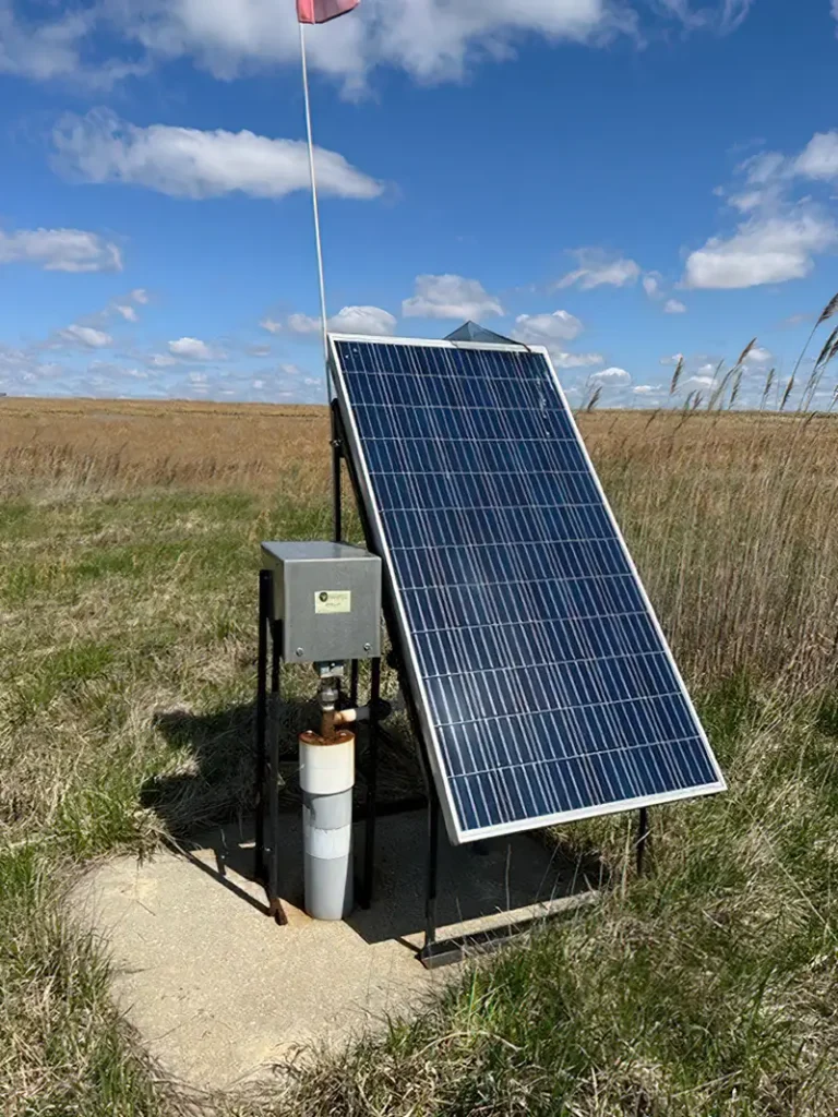 A solar panel is mounted on a metal frame in an open field with tall grass, connected to an electrical box and a cylindrical pipe below, resembling a top-head drive pump. A flag is attached to the top of the frame. The sky is partly cloudy with bright sunlight illuminating the scene.
