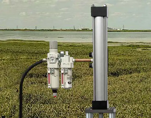 A close-up view of industrial equipment with cylindrical metallic components and attached gauges, featuring a top-head drive pump, set against an open grassy field and sky. The equipment appears to be mounted on a stand and connected by a black hose.