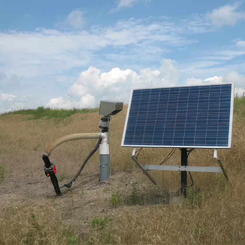A solar-powered top-head drive pump in a grassy area under a blue sky with scattered clouds. The setup includes a solar panel mounted on a metal frame and connected via hoses and pipes to the pump system anchored in the ground.