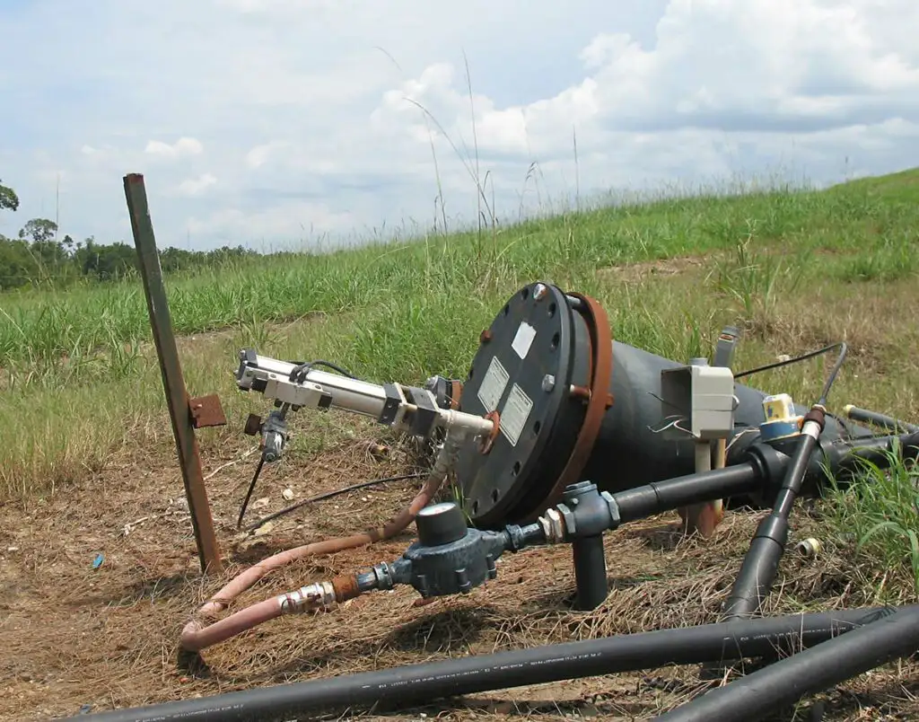 An outdoor setup of a water monitoring station on a grassy hillside features various pipes, valves, and a large black cylindrical device with an indicating gauge, complemented by a top-head drive pump. The sky is partly cloudy, and vegetation is visible in the background.